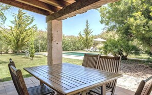 An outdoor patio with a wooden table and chairs, a concrete pillar, and a view of a swimming pool and trees. - Country House 'Cadernera' with Private Pool, Wi-Fi and Air Conditioning (Búger)