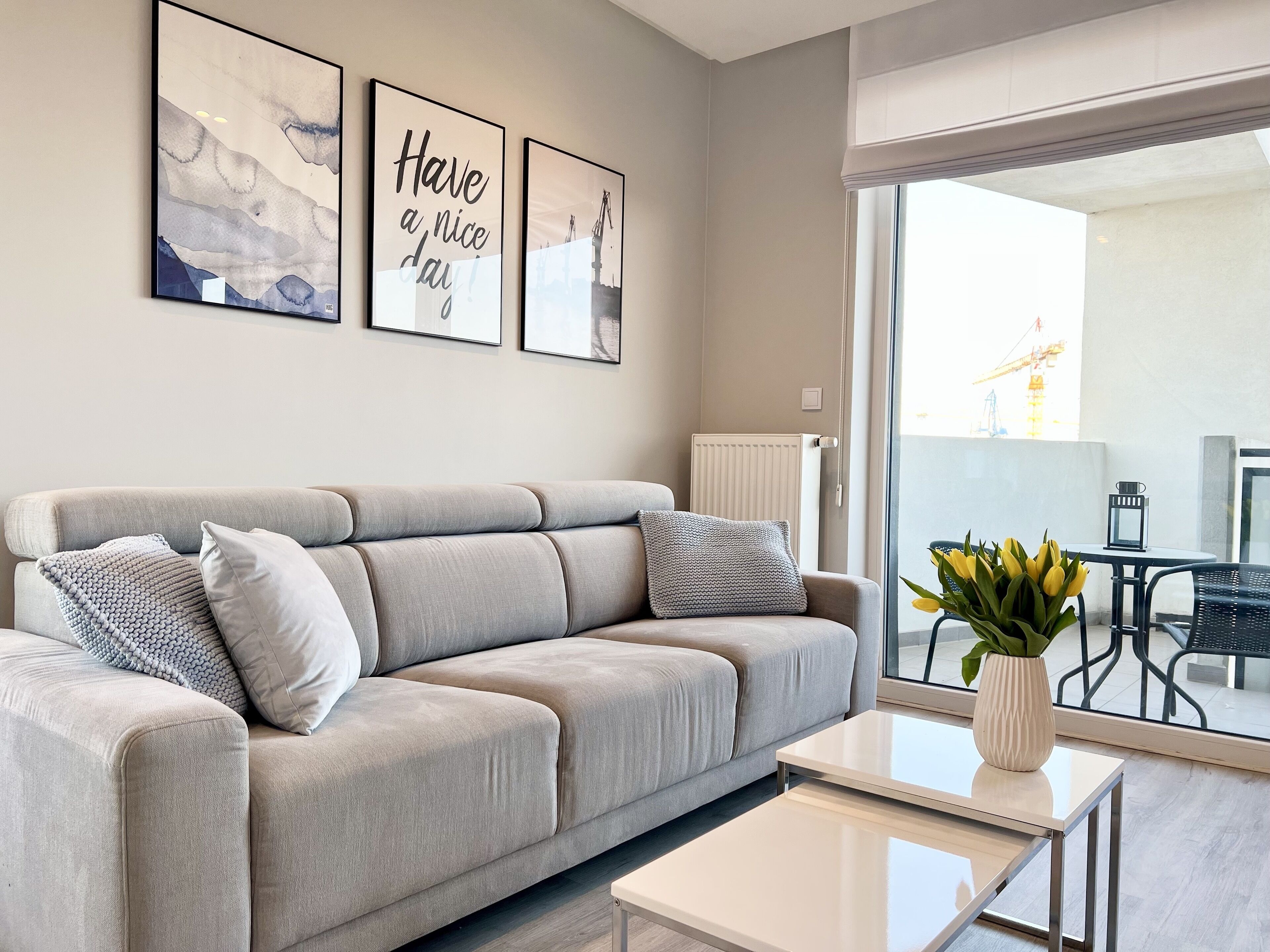 Ein modernes Wohnzimmer mit grauem Sofa, einem weißen Couchtisch und Blick auf einen Außenbalkon mit Tisch und Stühlen.