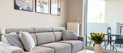 Ein modernes Wohnzimmer mit grauem Sofa, einem weißen Couchtisch und Blick auf einen Außenbalkon mit Tisch und Stühlen.