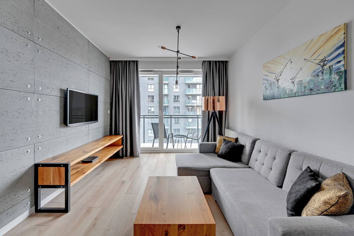 Una sala de estar moderna con un sofá gris, una mesa de centro de madera y un televisor montado en una pared con textura.