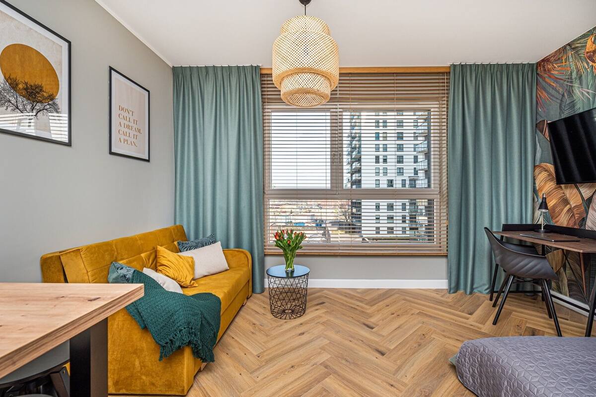 Una sala moderna con un sofá color mostaza, piso de madera en patrón de espiga, un ventanal con persianas y una lámpara colgante de fibras naturales.