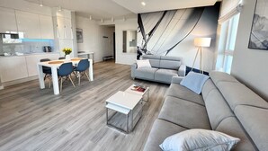 A modern living room with a grey sofa, a coffee table, and a dining area with a white table and blue chairs. - Bastion Wałowa by Q4Apartments (Gdansk)