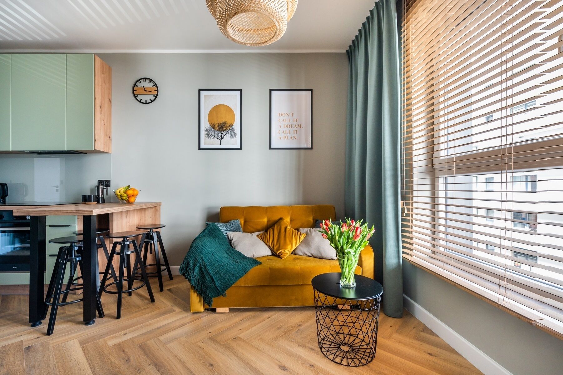 A modern living room with a yellow sofa, a coffee table, and a dining area with a wooden table and chairs. There are two framed pictures on the wall, a clock, and a window with blinds.