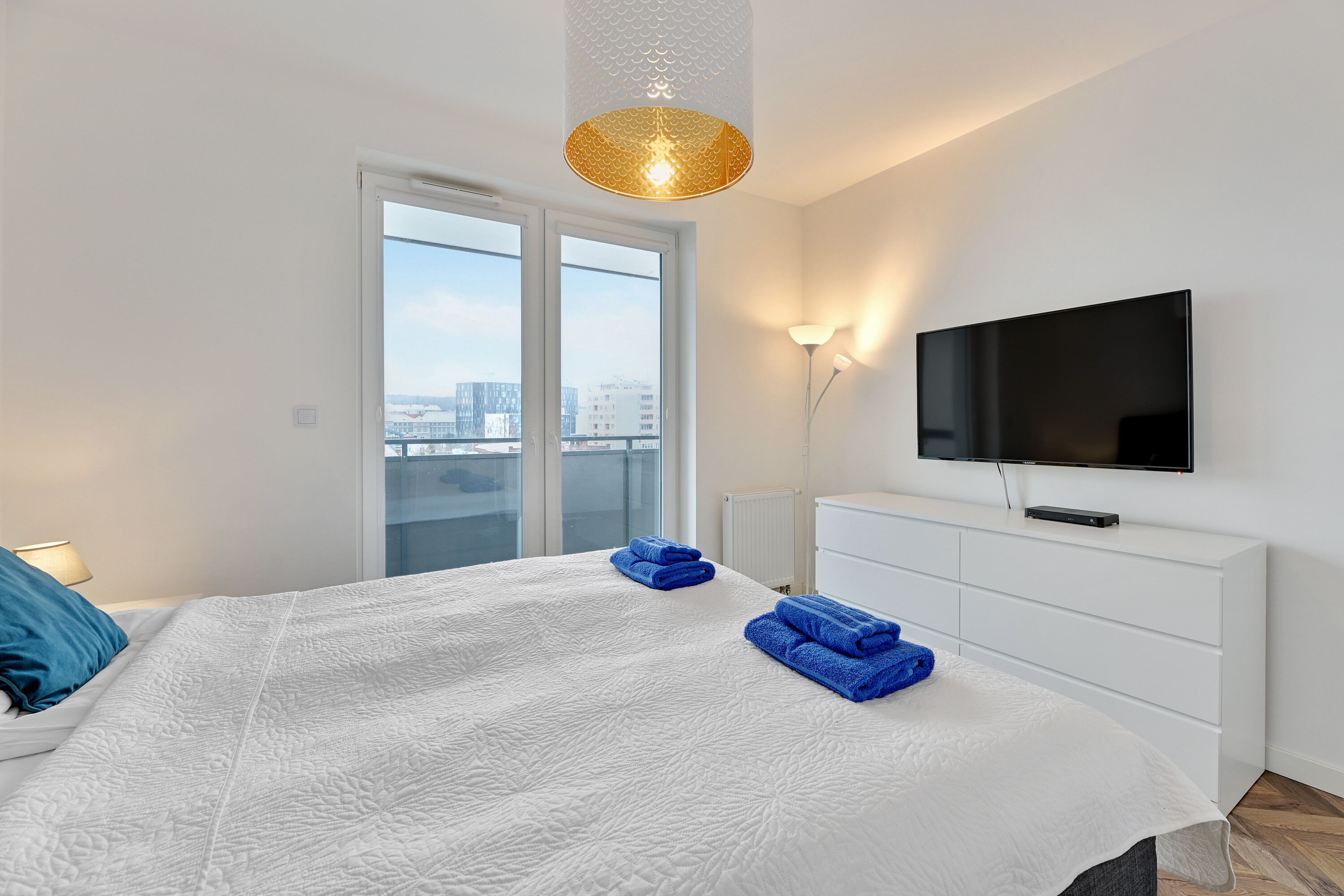 Ein modernes Hotelzimmer mit einem großen Bett, einem Flachbildfernseher, einem Balkon mit Blick auf die Stadt und einer weißen Kommode.