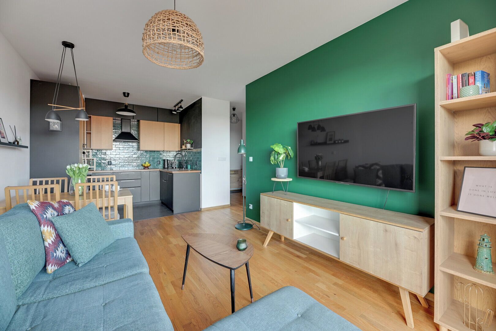 Ein modernes Wohnzimmer mit grüner Akzentwand, einem großen Flachbildfernseher, einem Holzesstisch und einem blauen Sofa.
