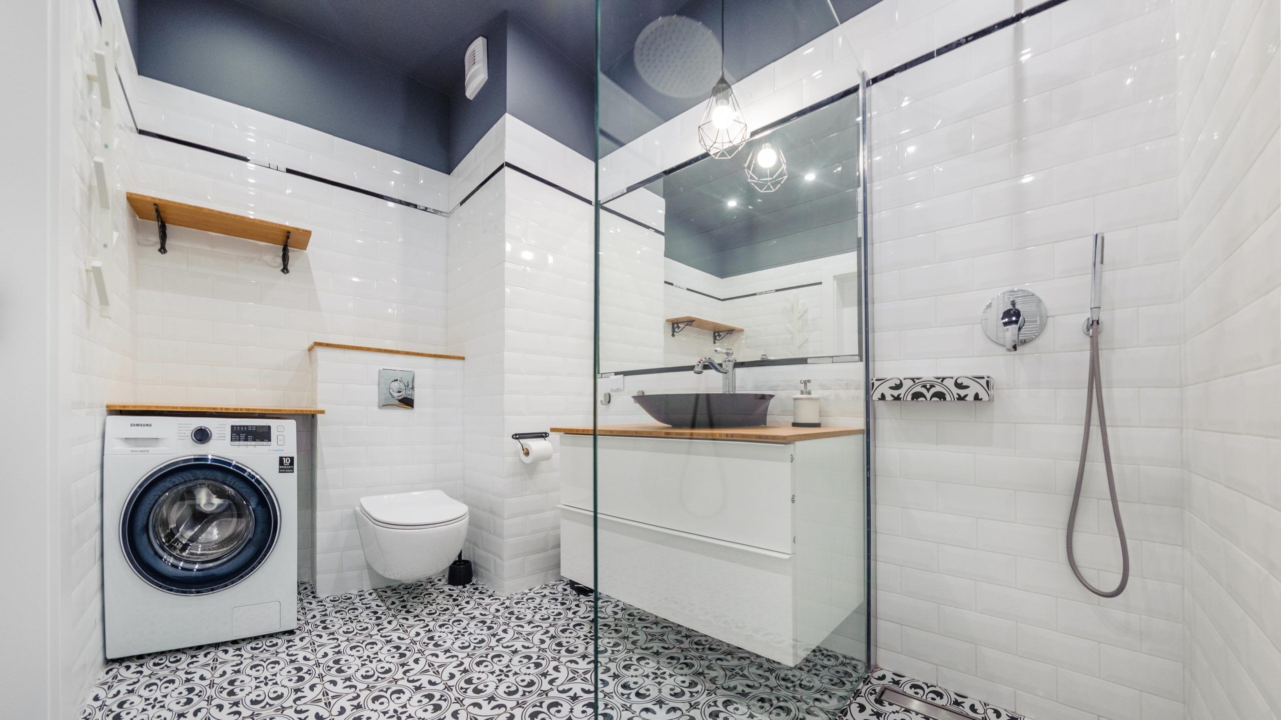 A modern bathroom with a washing machine, toilet, glass shower enclosure, and patterned floor tiles.