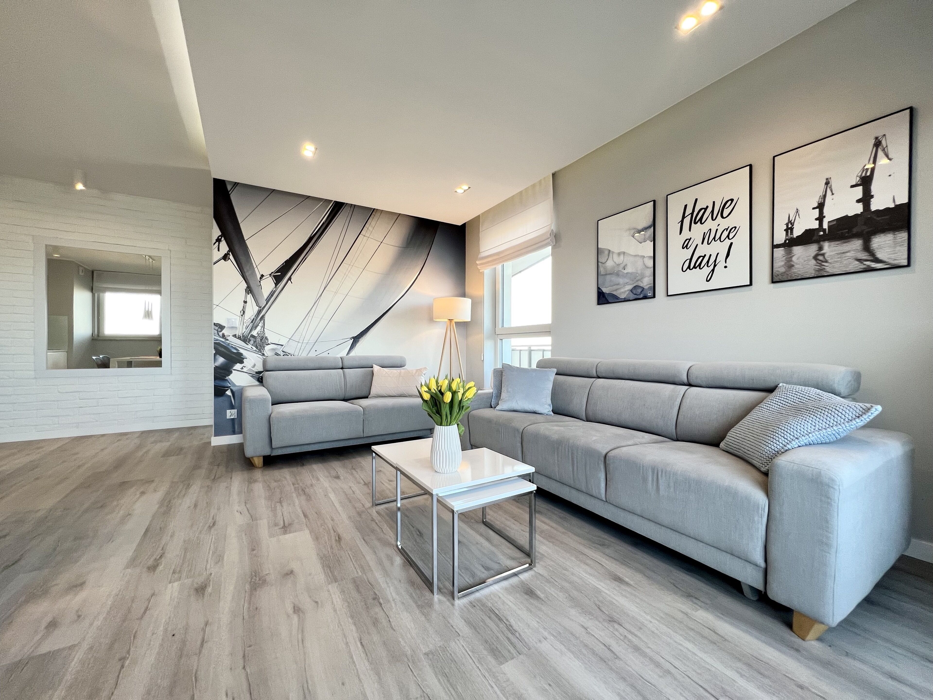 A modern living room with a grey sofa, a coffee table, and a hammock. There are framed pictures on the wall and a window providing natural light.
