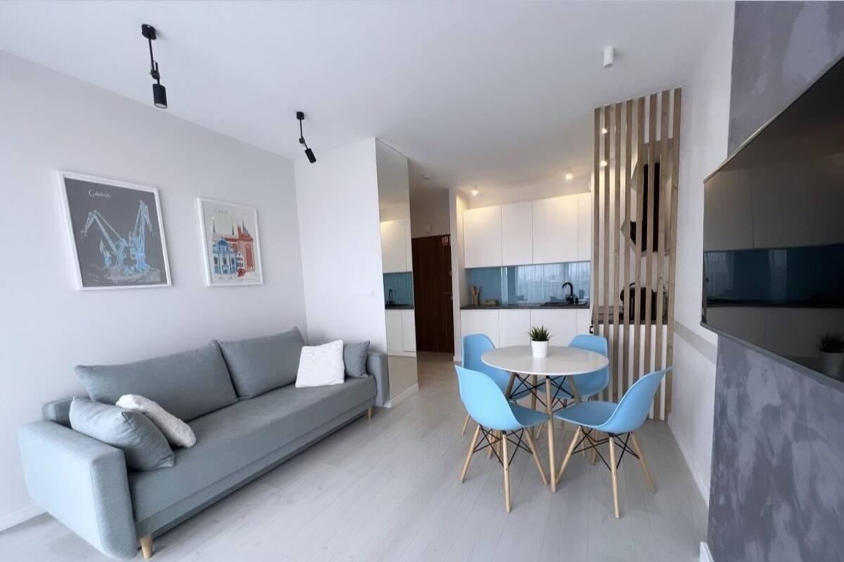 Una sala de estar moderna con un sofá gris, una mesa redonda con sillas azules y una cocina al fondo.