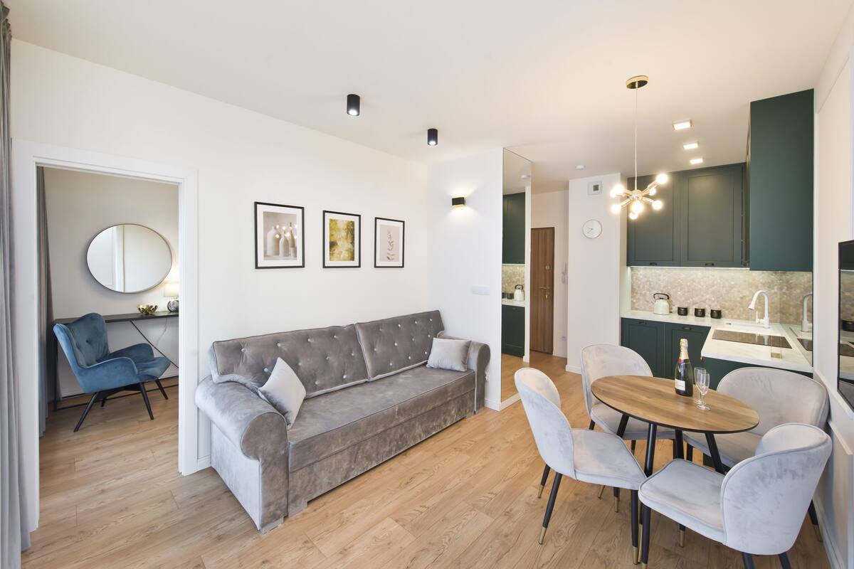 Una sala de estar moderna con un sofá gris, una silla azul y una mesa de comedor de madera con cuatro sillas. Se observa una cocina con armarios verdes y una encimera blanca.