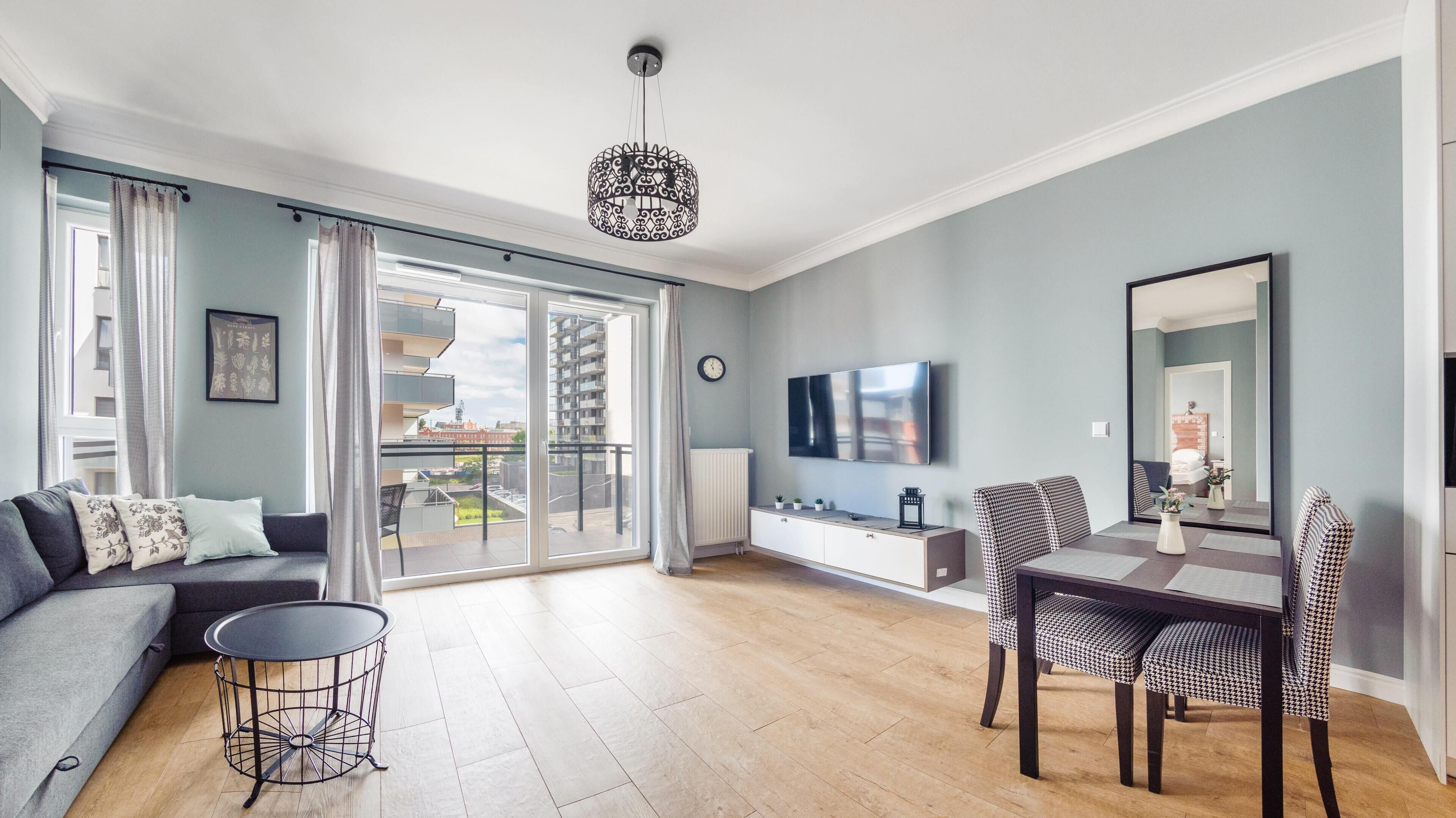 A modern living room with a grey sofa, a dining table with chairs, a TV, and a balcony with a view of buildings.
