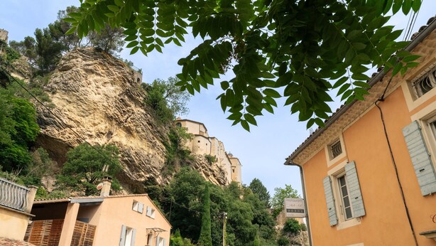 Um edifício na encosta de uma colina, com um precipício íngreme, uma varanda e uma rua com edifícios.