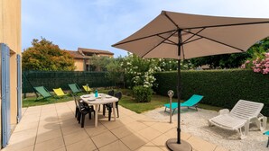 Um pátio externo com mesa, cadeiras, um grande guarda-sol e espreguiçadeiras.