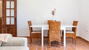 Un comedor con una mesa blanca y sillas de mimbre, un jarrón con flores secas y una puerta de madera.