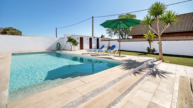 Una piscina con agua cristalina, rodeada por un muro blanco y un patio empinado con sillas de descanso y una sombrilla verde.