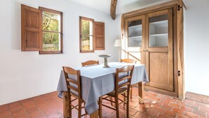 A dining area with a table set for four, wooden chairs, a window with open shutters, and a wooden cabinet with glass doors. - Villa 'Casa Di Pruno' with Private Pool, Wi-Fi and Air Conditioning (Figari)