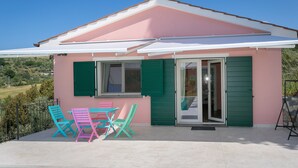 A small house with a pink exterior, green shutters, and a white awning. There are two chairs and a table outside. - Holiday Home 'Casetta Maya' with Sea View, Wi-Fi and Air Conditioning (Riparbella)