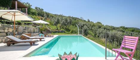 A pool with lounge chairs and a pink chair, surrounded by greenery and a clear sky.