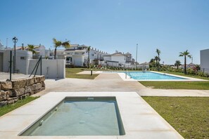 Un espace piscine avec un jacuzzi, entouré d’une clôture, et un bâtiment à la façade blanche en arrière-plan.