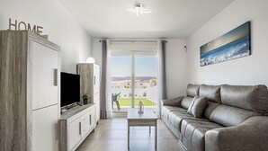 A modern living room with a sofa, TV, and a table. There's a balcony with a view. - Apartment 'Apartamento Poniente' with Sea View, Wi-Fi and Air Conditioning (Zahara de los Atunes)