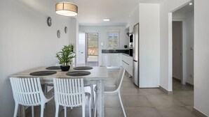 A modern kitchen with white cabinetry, a dining area with a wooden table and white chairs, and a view through the window. - Apartment 'Apartamento Poniente' with Sea View, Wi-Fi and Air Conditioning (Zahara de los Atunes)