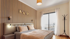 Une chambre à coucher comprenant un lit, des tables de chevet, un luminaire fixé au mur, un porte-manteau et une grande fenêtre avec des rideaux.