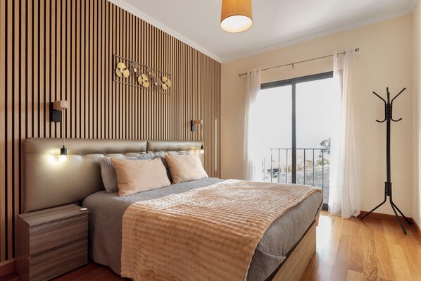 Une chambre à coucher comprenant un lit, des tables de chevet, un luminaire fixé au mur, un porte-manteau et une grande fenêtre avec des rideaux.