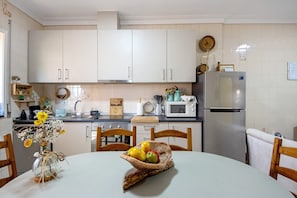 A kitchen with white cabinets, a microwave, a refrigerator, a wooden table with a fruit bowl, and a vase with flowers.