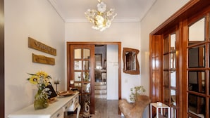 A hallway with a wooden sign, a chandelier, a white dresser, a vase with flowers, and a patterned rug.