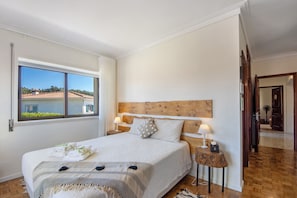 A bedroom with a large bed, two bedside tables with lamps, a wooden headboard, and a window with a view of buildings.