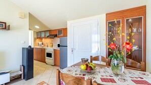 Un coin repas avec une table dressée pour deux personnes, un vase rempli de fleurs et, au fond, une cuisine.