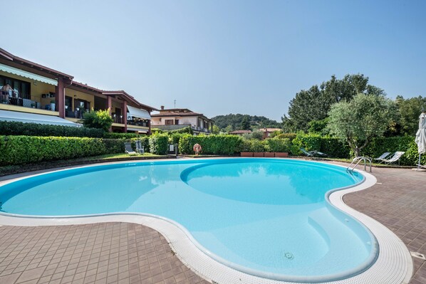 A swimming pool with a tiled deck, surrounded by greenery and buildings.