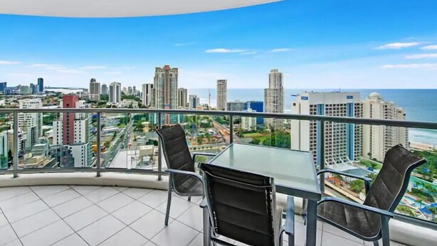 A balcony with a glass table and chairs, overlooking a cityscape and the sea.