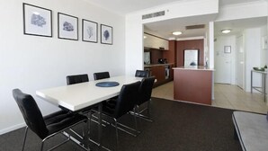 A modern dining area with a white table, black chairs, and a small island with a refrigerator. There are framed pictures on the wall and a kitchen in the background.
