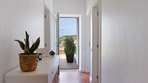 Un couloir avec une plante en pot posée sur une console blanche, qui mène à une porte ouverte donnant sur un balcon et sur lequel on aperçoit une grande plante en pot à l’extérieur.