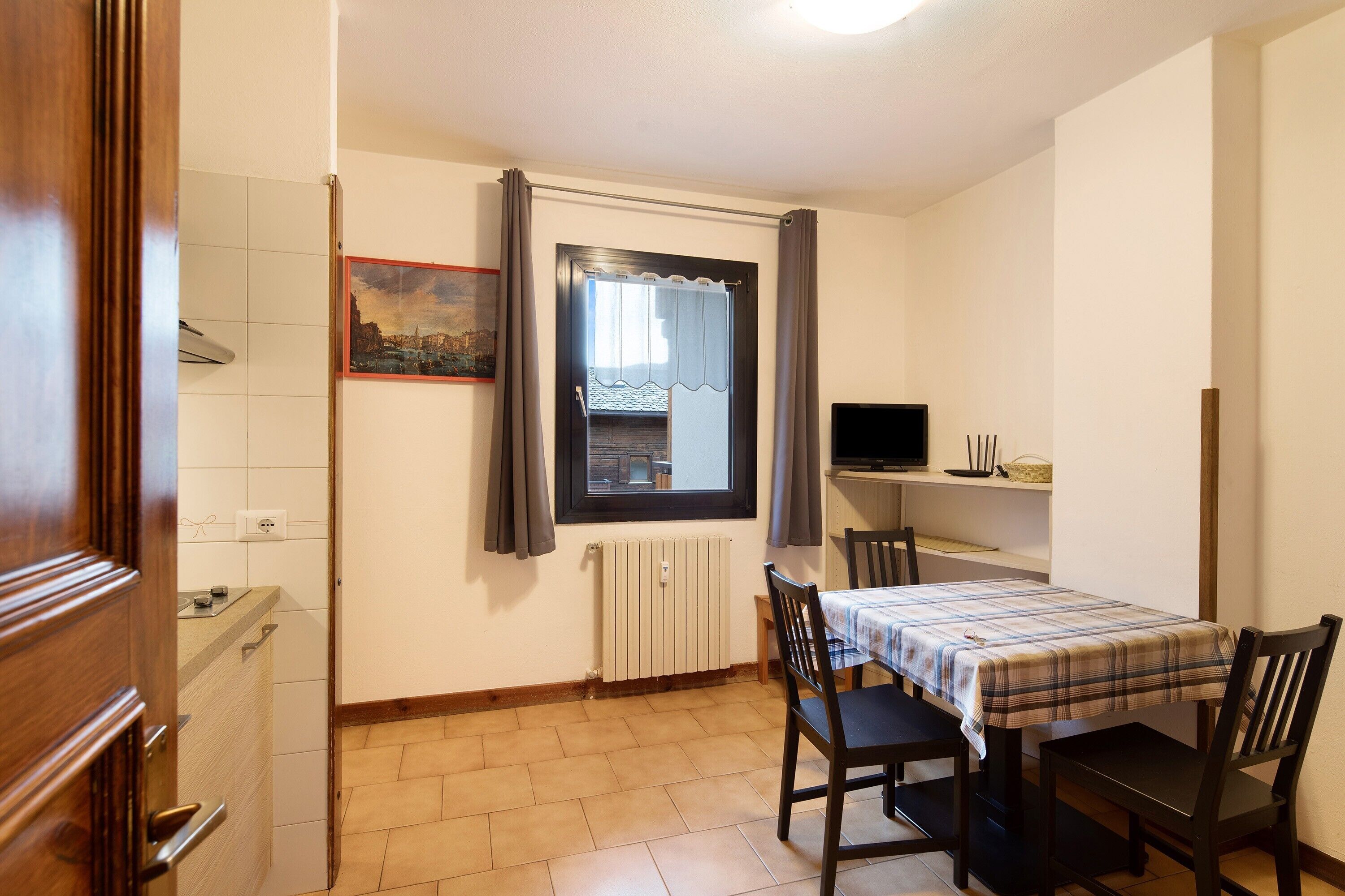 A small room with a dining table, chairs, a window with curtains, a radiator, and a television on a shelf.