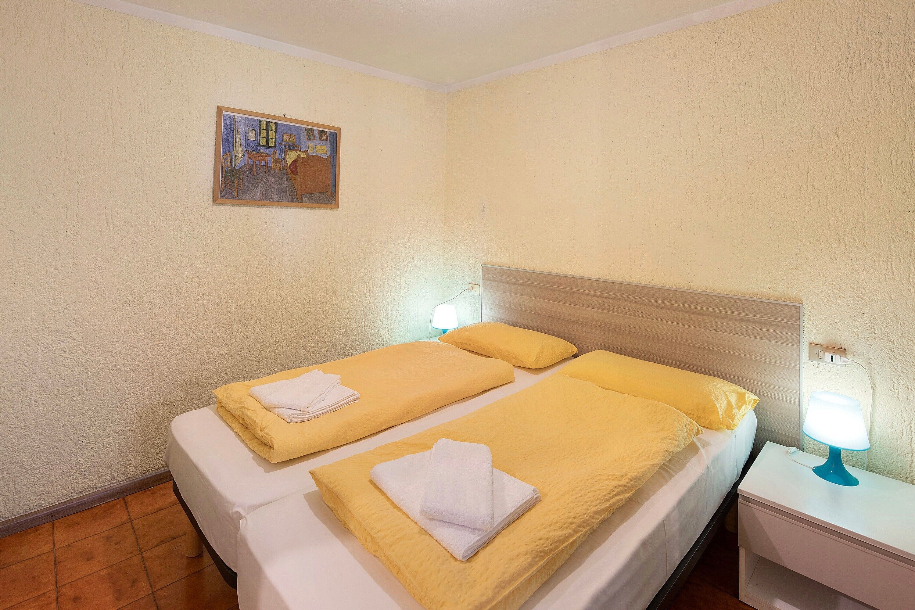 A hotel room with two beds, each with a yellow bedspread and white towels, a wooden headboard, a nightstand with a lamp, and a painting on the wall.