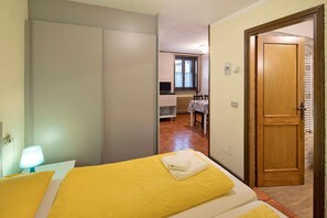 A bedroom with a bed, a bedside lamp, a wooden door, and a view into a kitchen area.