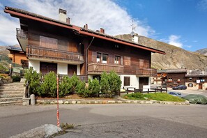 A two-story wooden house with balconies, a stone staircase, and a mountain backdrop.