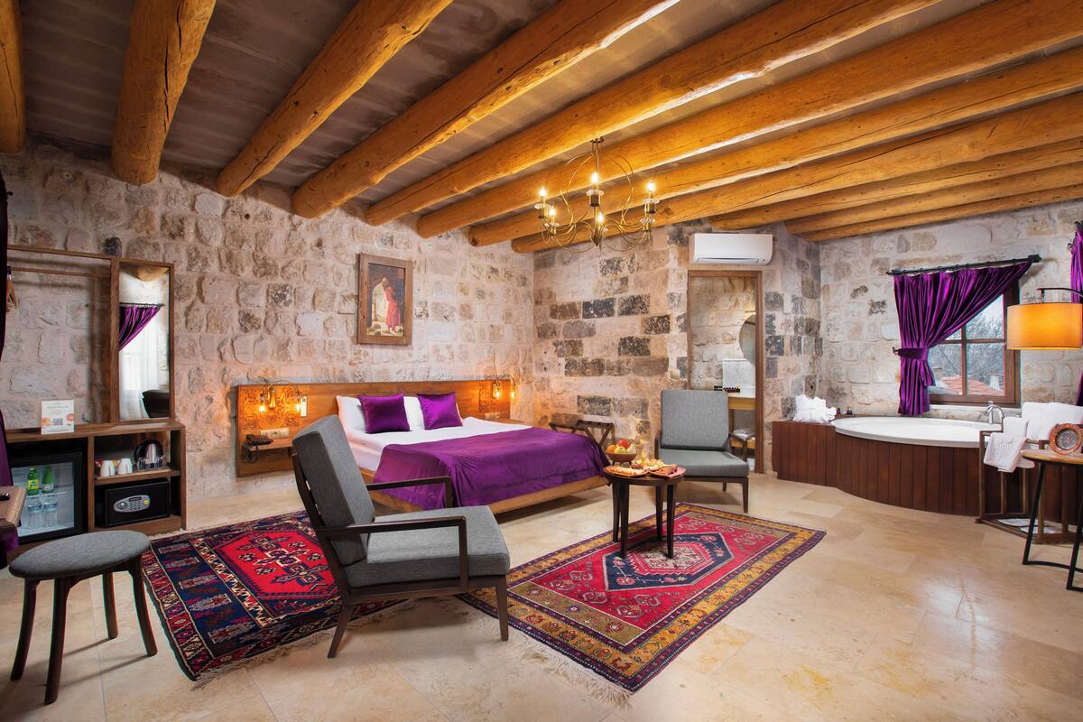 A rustic room with a stone wall, wooden beams, a bed with purple bedding, a sitting area with a chair and table, and a bathtub.