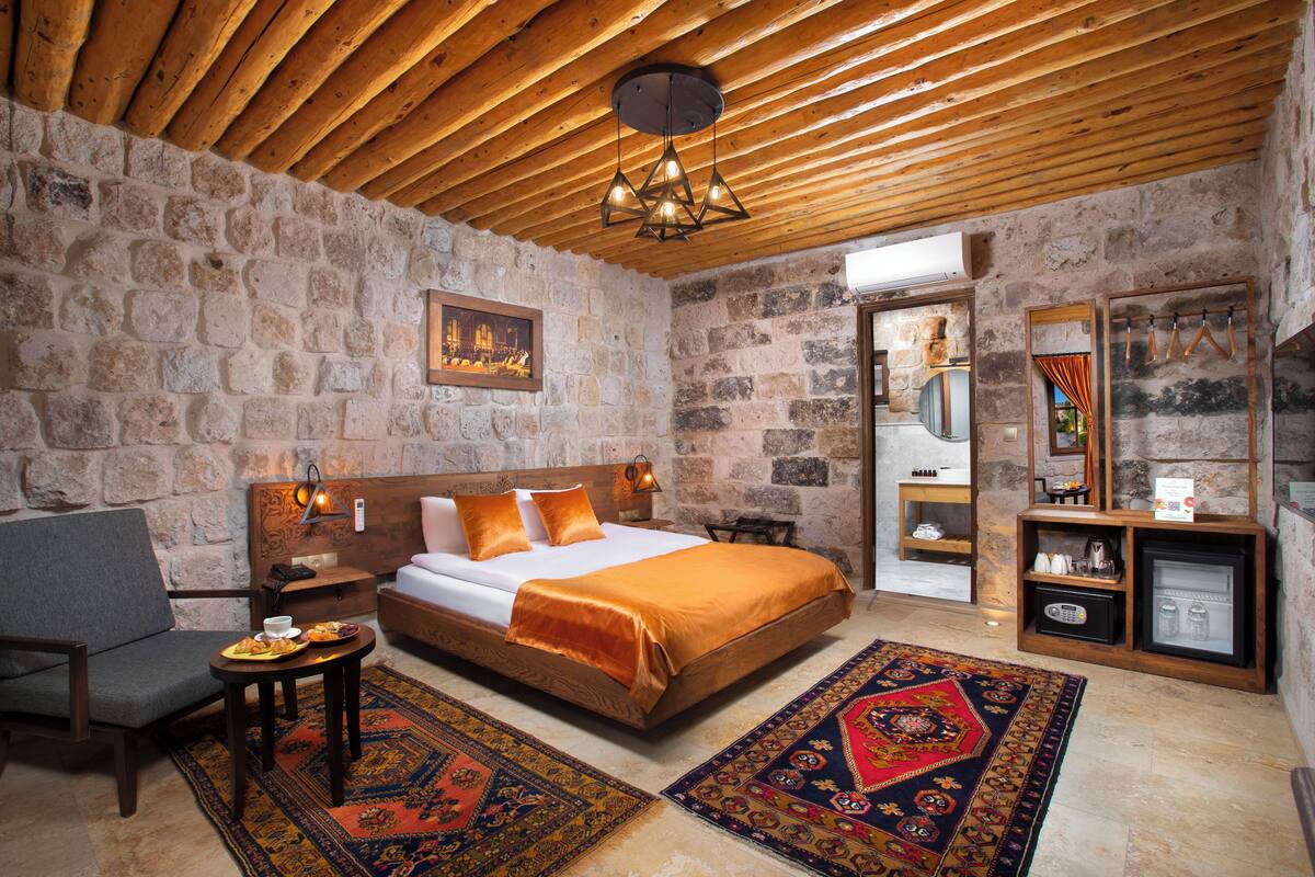 A cozy room with a stone wall, a wooden ceiling, a bed with an orange blanket, a grey chair, a small table with a tray of food, and a decorative rug.