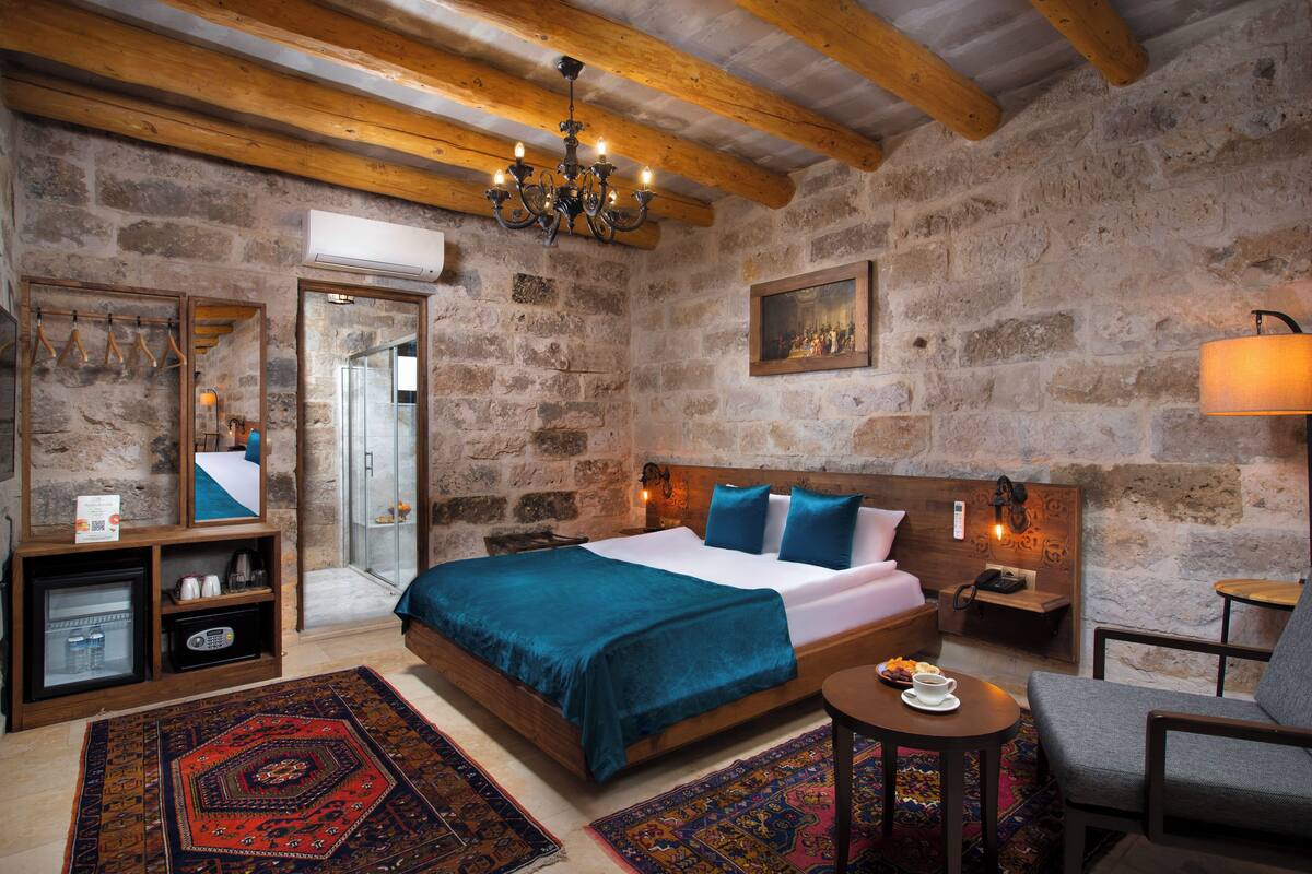 A rustic room with a stone wall, wooden beams, a bed with blue bedding, a small table with a cup, and a chair.