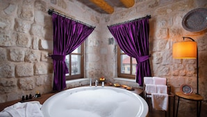 A stone-walled bathroom with a large bathtub, purple curtains, a wooden bench with towels, and a small table with a lamp. - Woox Cappadocia Hotel (Nevsehir)