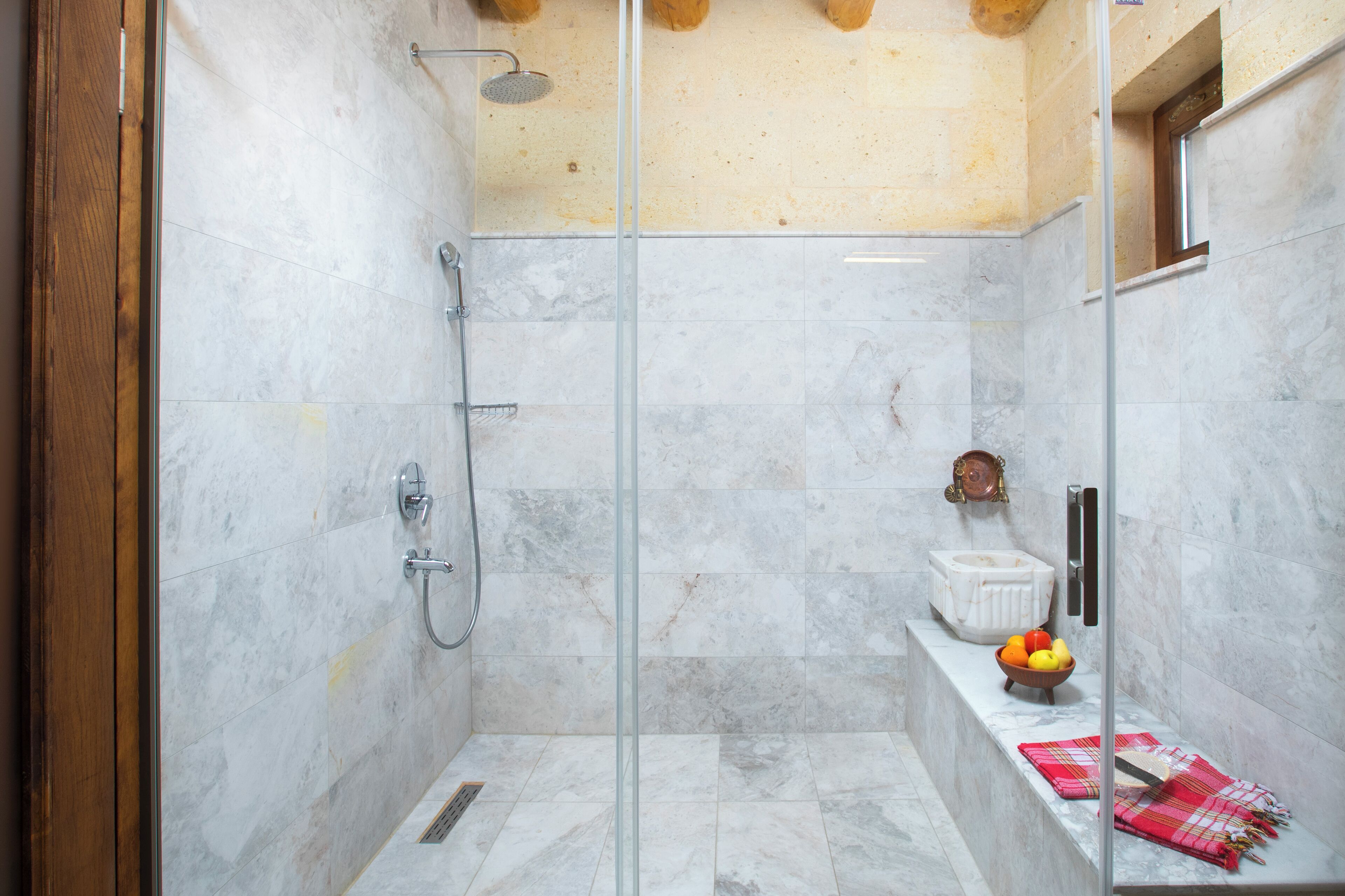 A modern shower with glass doors, a marble-tiled shower area, and a small bench with a bowl of fruit and a towel.