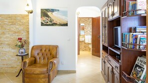 A living room with a leather armchair, a bookshelf, a wall-mounted lamp, a framed picture of a beach, and a doorway leading to a bathroom. - Vacation Home "Bela Casa Na Costa Vicentina" with Private Pool, Private Garden & Wi-Fi (Aljezur)