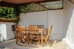 A wooden table and chairs set up outdoors, with a white wall and a wooden trellis covered in foliage in the background.