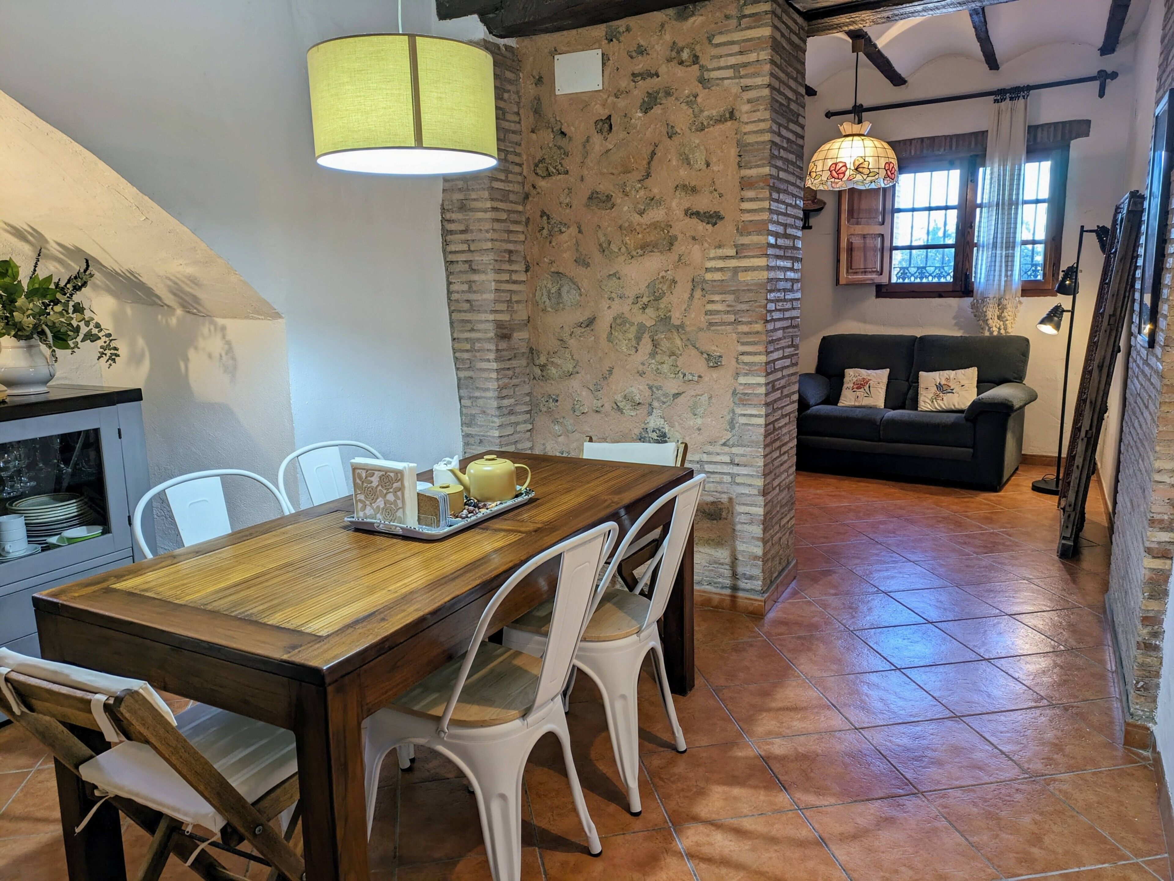 A dining area with a wooden table, white chairs, a yellow lamp, and a stone wall.