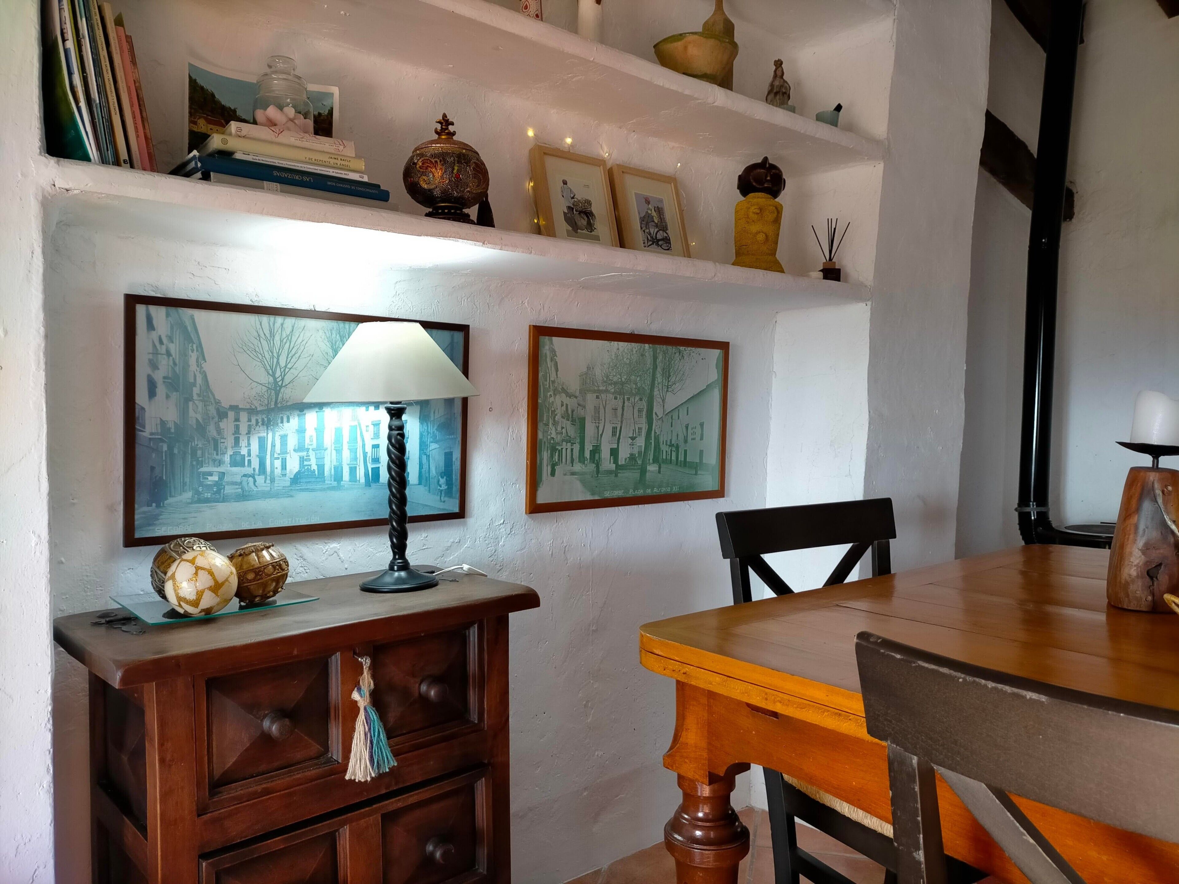 Een eethoek met een houten tafel, stoelen, een lamp, een kast en wandplanken met diverse spullen.