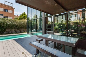 A modern outdoor pool area with glass walls, wooden deck, and outdoor furniture.