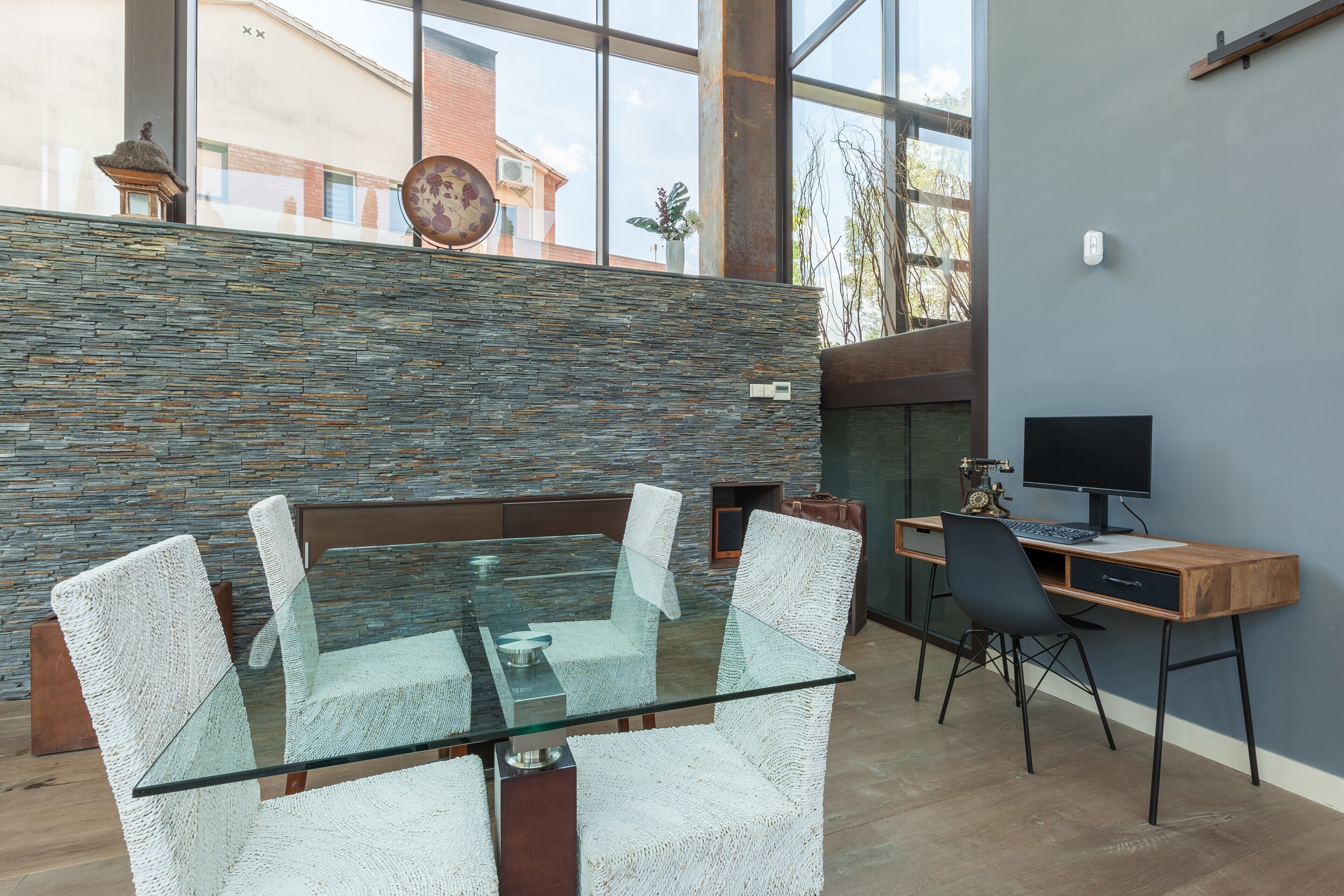 Un interior moderno con una mesa de vidrio, sillas tejidas, una pared de piedra, una chimenea y un escritorio con ordenador.