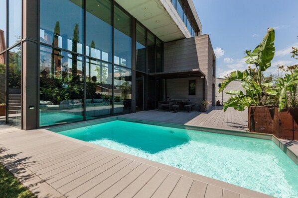 A modern outdoor pool area with a wooden deck, large glass windows, and a patio with outdoor furniture.